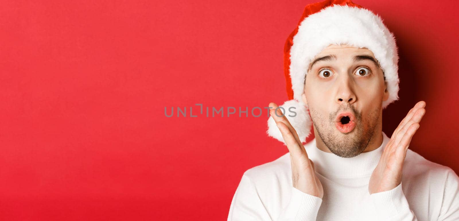 Close-up of amazed handsome man in santa hat and white sweater, raising hands and looking impressed, standing over red background by Benzoix