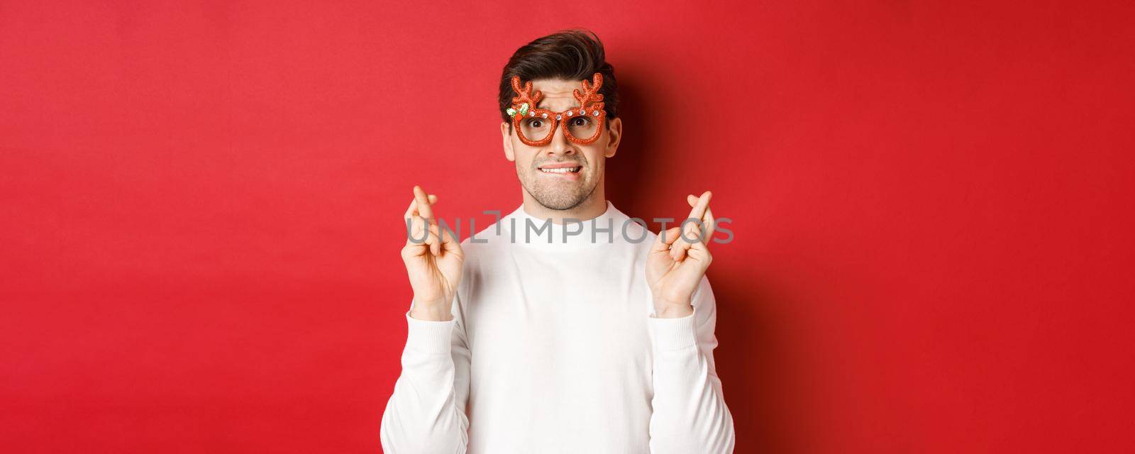 Close-up of handsome nervous guy in party glasses, making a wish, crossing fingers for good luck and looking with hope at camera, standing over red background.