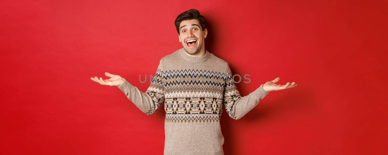 Portrait of happy and surprised, handsome caucasian guy, wearing christmas sweater, spread hands sideways and looking clueless, standing over red background.