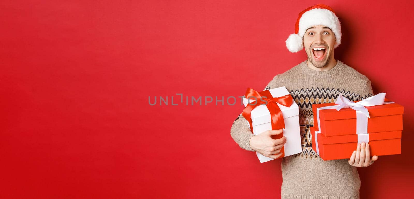 Concept of winter holidays, new year and celebration. Image of amazed and happy man in sweater and santa hat, holding christmas gifts and shouting for joy, red background by Benzoix