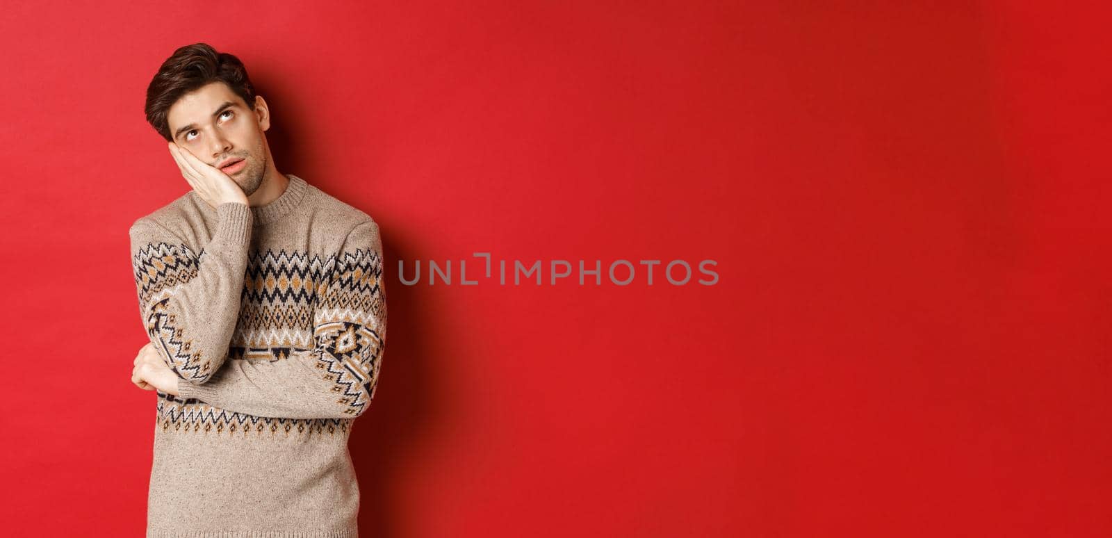 Image of annoyed young man in christmas sweater, roll eyes and leaning on hand, express displeasure and irritation, standing over red background.