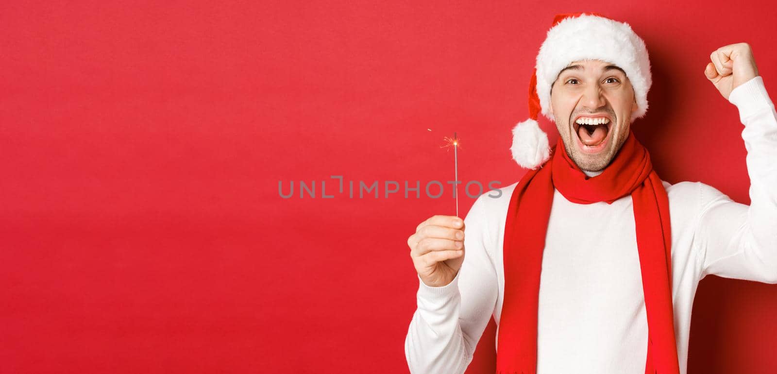 Concept of christmas, winter holidays and celebration. Portrait of excited handsome man, raising hand up and holding sparkler, wishing happy new year, standing over red background by Benzoix