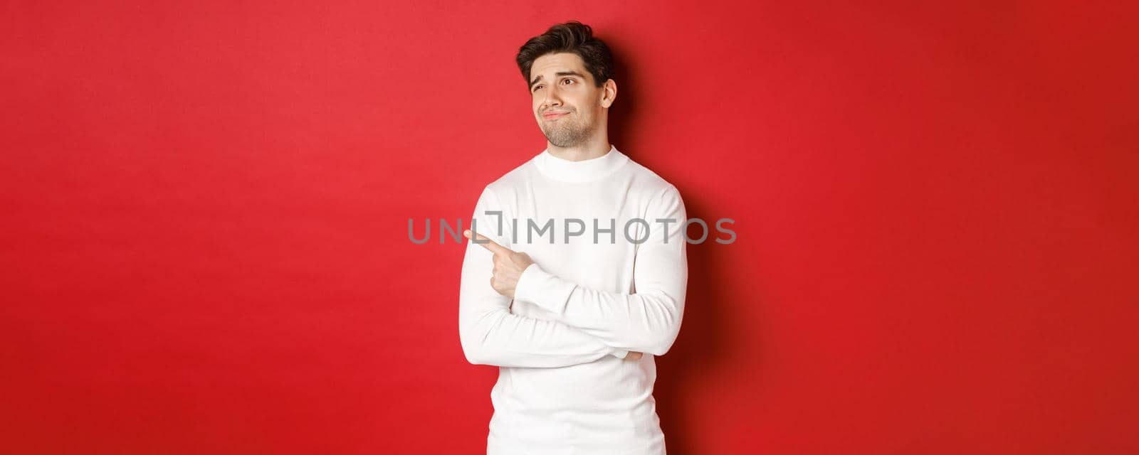 Concept of winter holidays. Image of displeased young man in white sweater, looking and pointing finger left at something average, standing skeptical over red background.