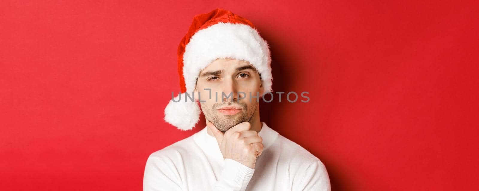 Close-up of thoughtful handsome man in santa hat, frowning and looking at camera, thinking about something, standing over red background by Benzoix