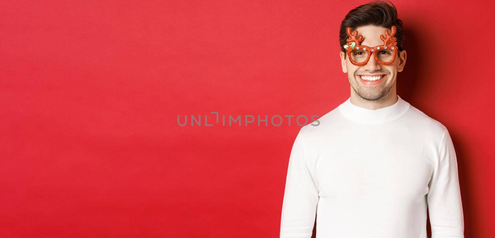 Close-up of attractive man in white sweater and party glasses, celebrating christmas, smiling amused, standing over red background.