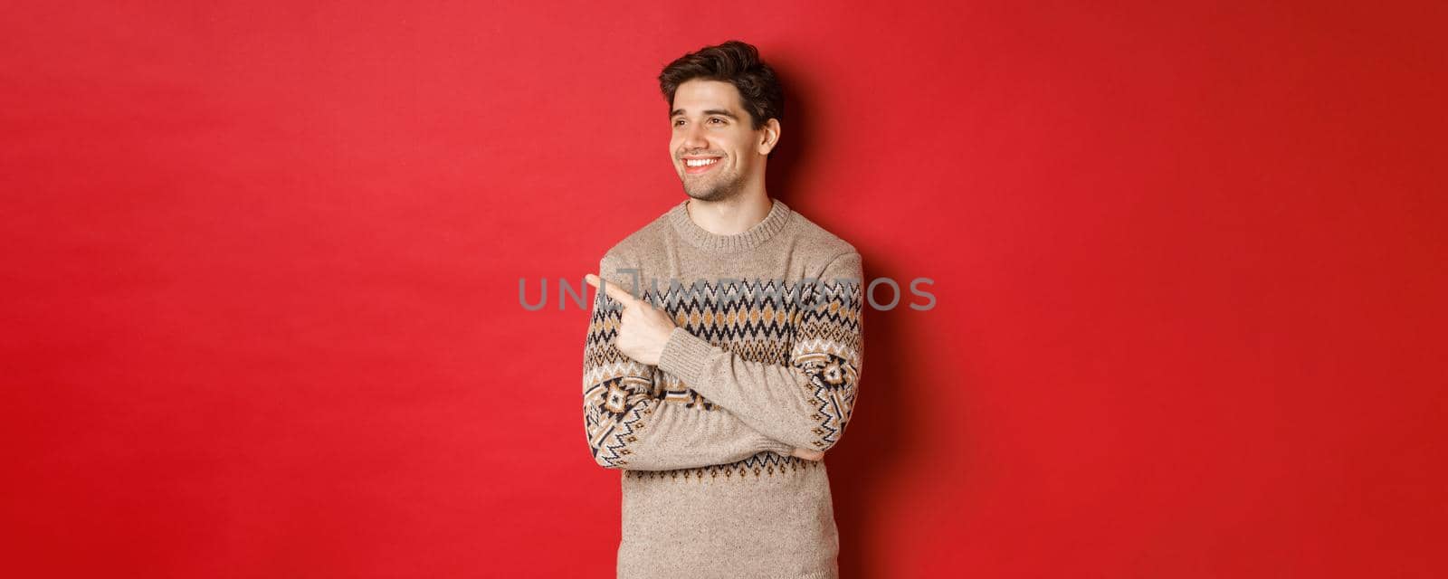 Portrait of attractive caucasian man celebrating new year, winter holidays, pointing finger and looking left with pleased smile, showing christmas advertisement, standing over red background by Benzoix