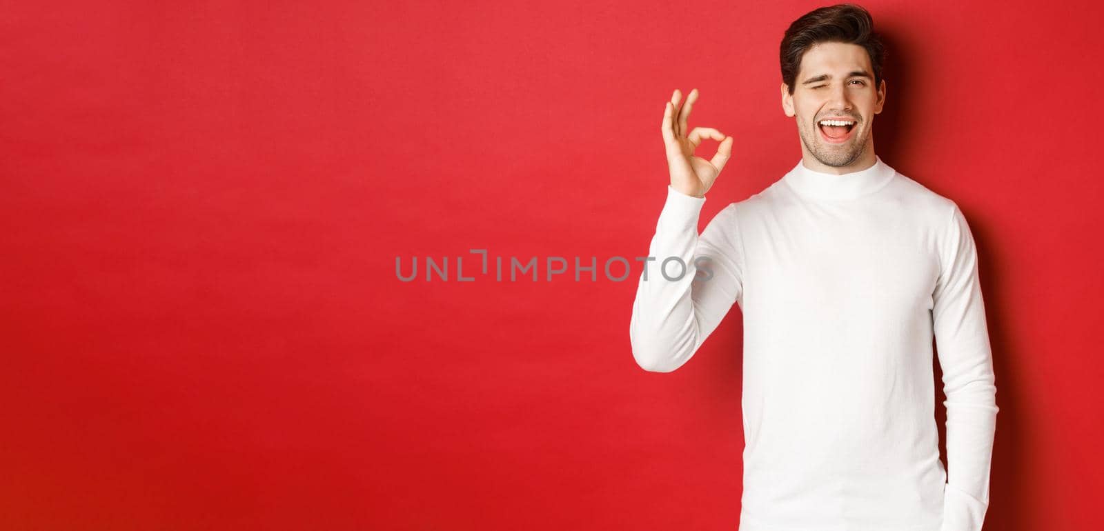Concept of winter holidays, christmas and lifestyle. Handsome man in white sweater winking at you, showing okay sign and smiling, guarantee quality, recommending something by Benzoix