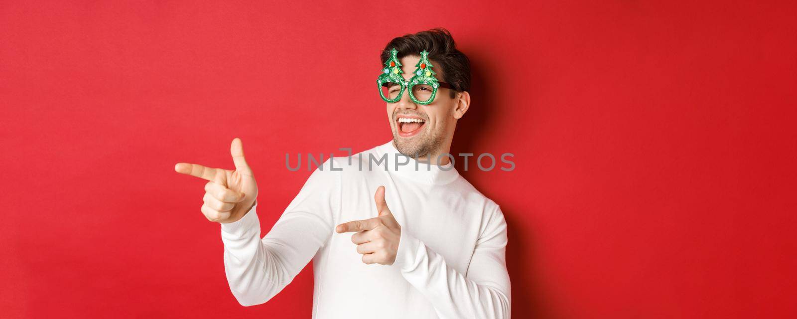 Close-up of cheeky handsome man in party glasses, pointing finger left and greeting person, wishing merry christmas, standing over red background by Benzoix