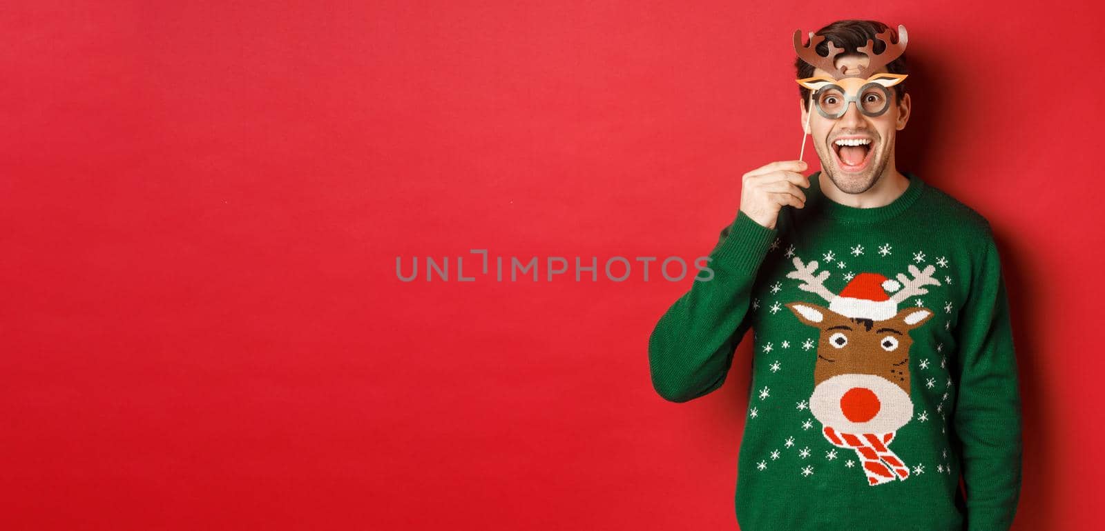 Handsome surprised man in christmas sweater, holding party mask and smiling, enjoying new year celebration, standing over red background by Benzoix