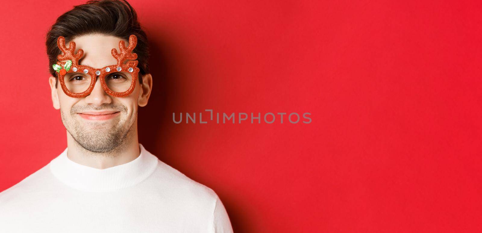 Concept of winter holidays, christmas and celebration. Close-up of attractive smiling male model in party glasses, standing against red background.
