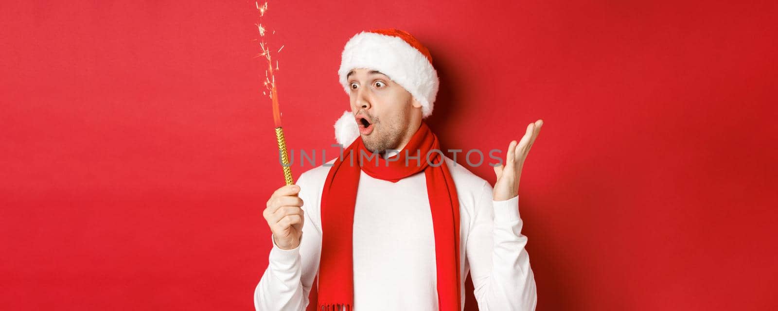 Concept of christmas, winter holidays and celebration. Close-up of amazed handsome man in santa hat and scarf, looking at sparkler with excitement, standing over red background by Benzoix