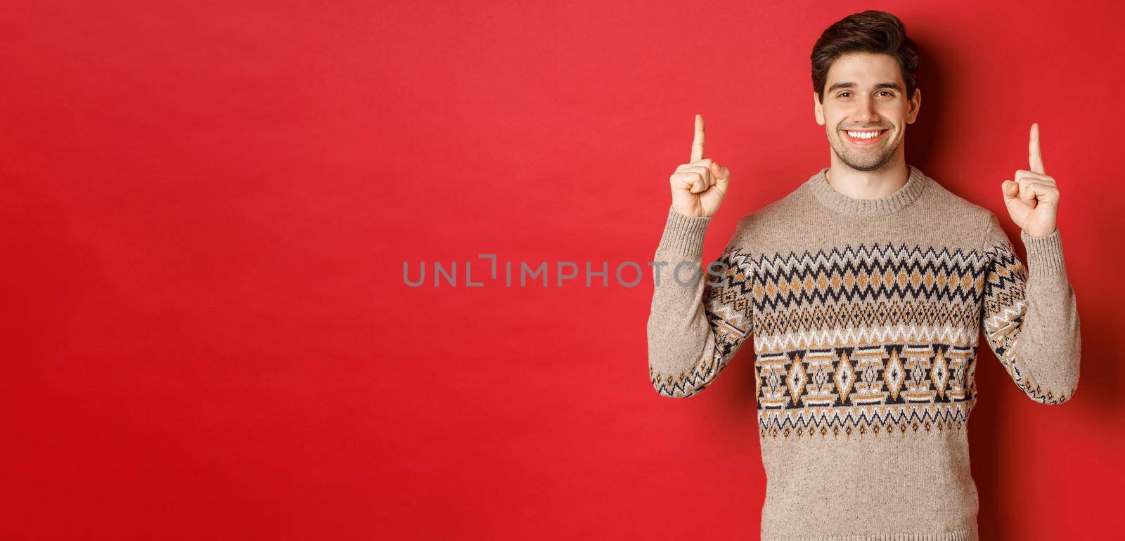 Portrait of attractive guy in christmas sweater pointing fingers up, smiling and showing winter holidays advertisement, celebrating new year over red background by Benzoix