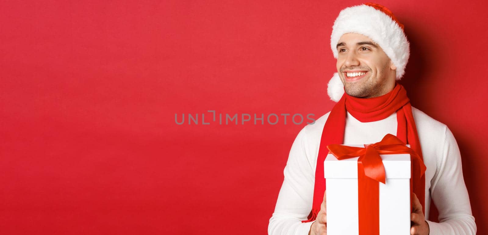 Concept of winter holidays, christmas and lifestyle. Close-up of attractive guy in santa hat and scarf, smiling and looking left while holding new year present, standing over red background.