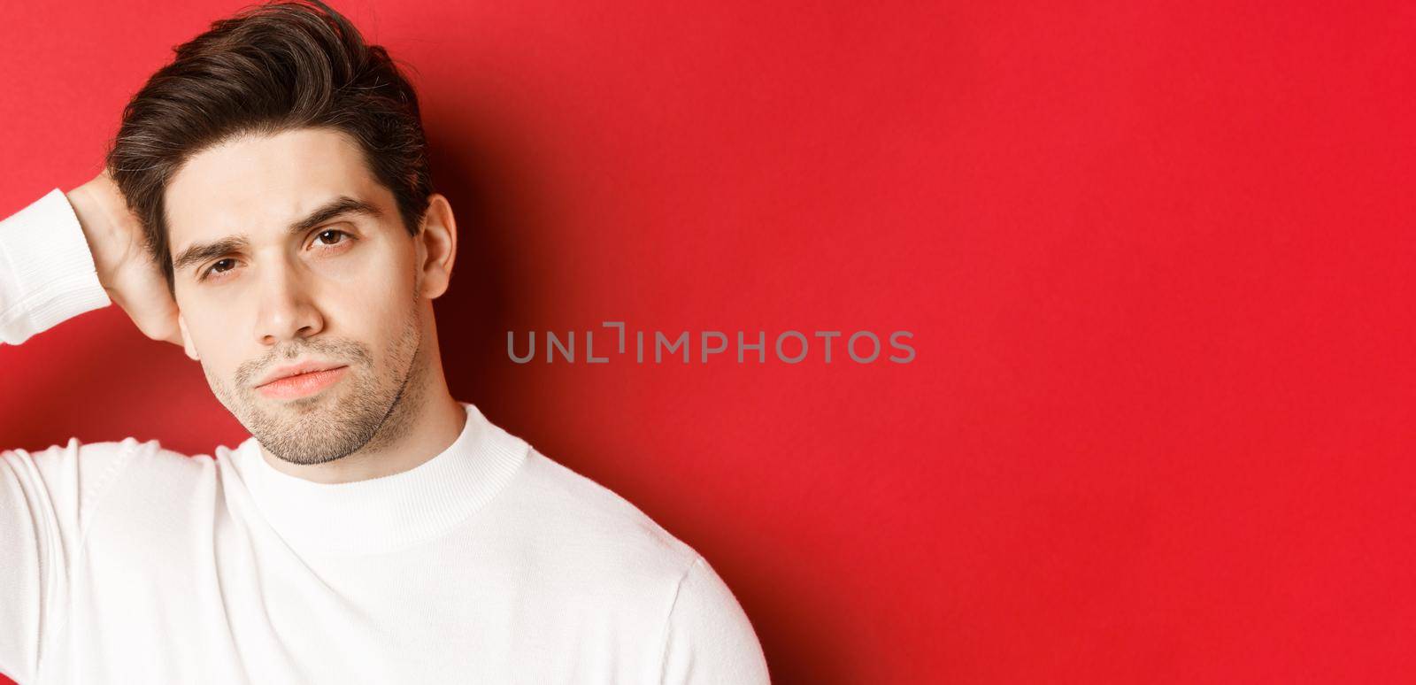 Close-up of handsome brunette man touching his haircut and looking sassy at camera, standing in white sweater over red background.