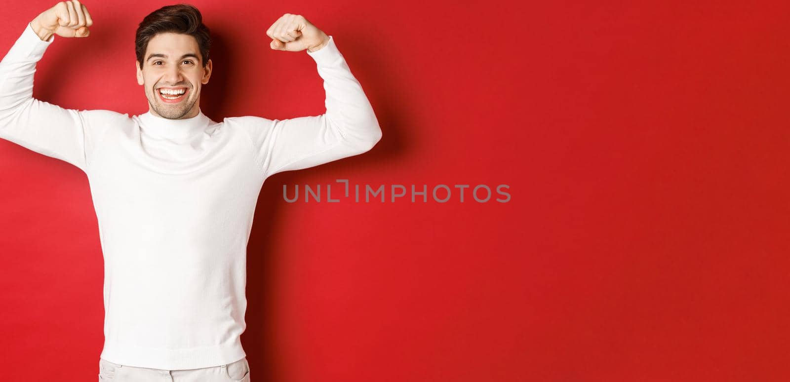 Portrait of smiling handsome man in white sweater, flexing biceps and bragging with strength, show-off strong muscles after workout, standing over red background by Benzoix