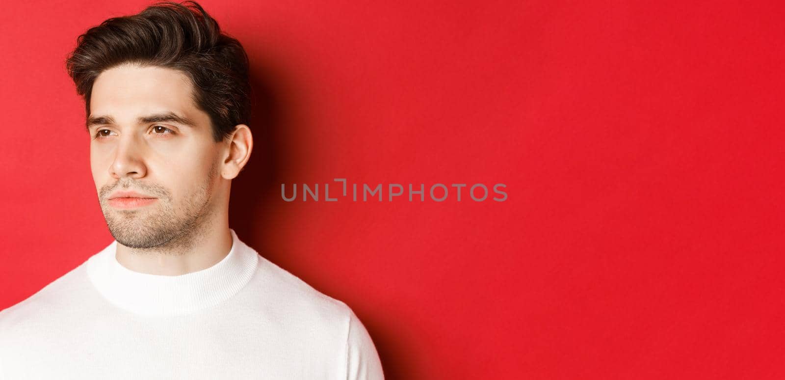 Close-up of serious and thoughtful man in white sweater, looking left, standing against red background.