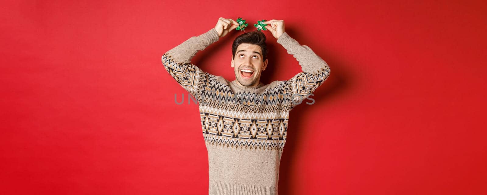 Concept of winter holidays, christmas and celebration. Image of funny and cute adult man enjoying new year party, smiling and rejoicing, standing over red background by Benzoix