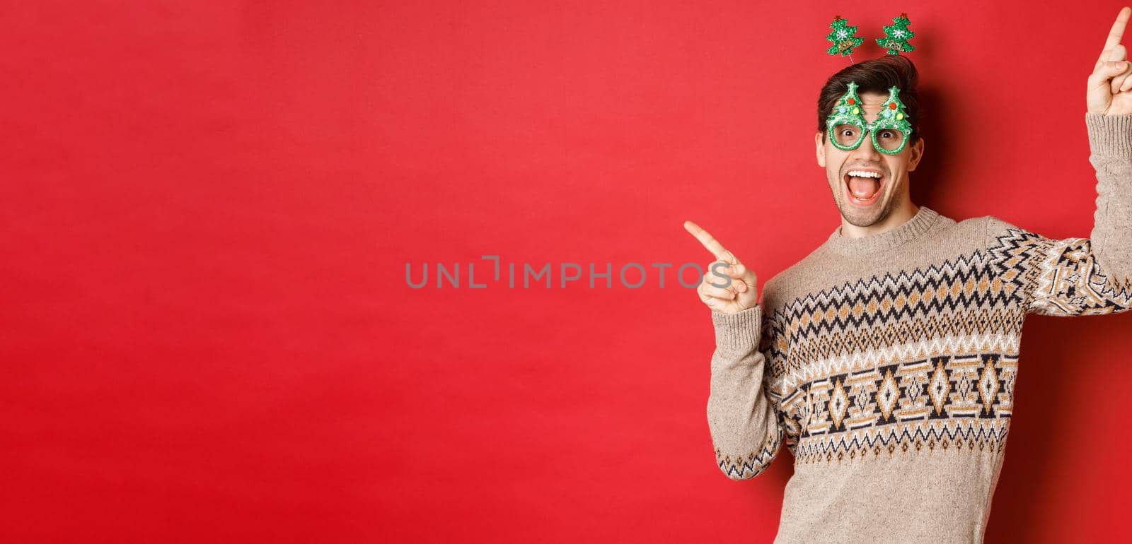 Portrait of joyful handsome guy in party glasses and christmas sweater, dancing and pointing fingers sideways, enjoying new year celebration, standing over red background by Benzoix