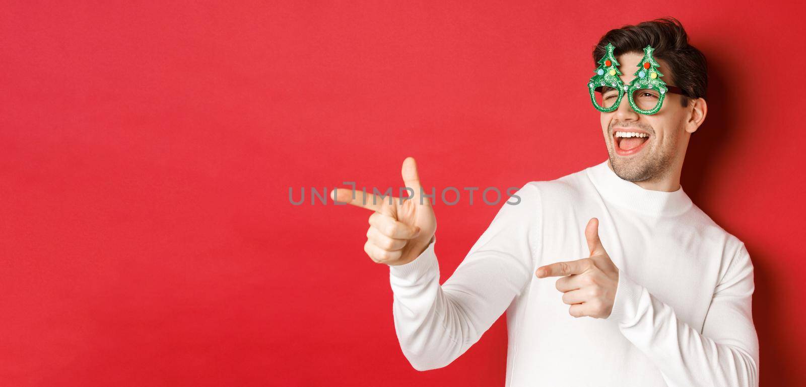 Close-up of cheeky handsome man in party glasses, pointing finger left and greeting person, wishing merry christmas, standing over red background by Benzoix