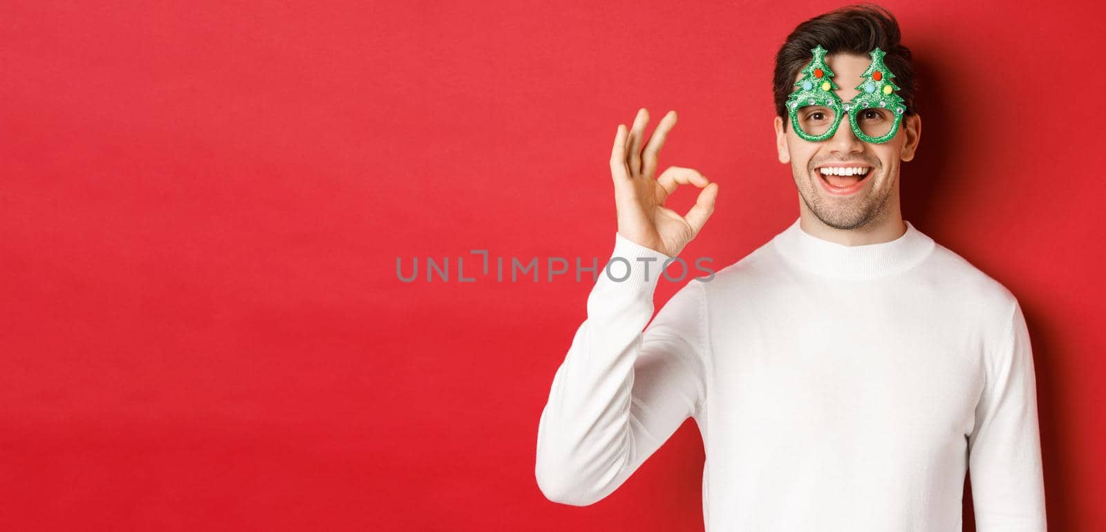 Happy caucasian guy wishing merry christmas, celebrating winter holidays, showing okay sign and smiling pleased, standing over red background.