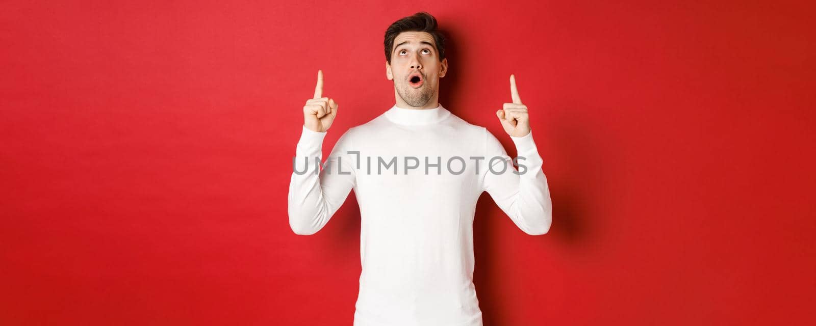 Concept of winter holidays. Portrait of handsome surprised man in white sweater, reacting to new year promo offer, looking and pointing fingers up, standing impressed against red background by Benzoix