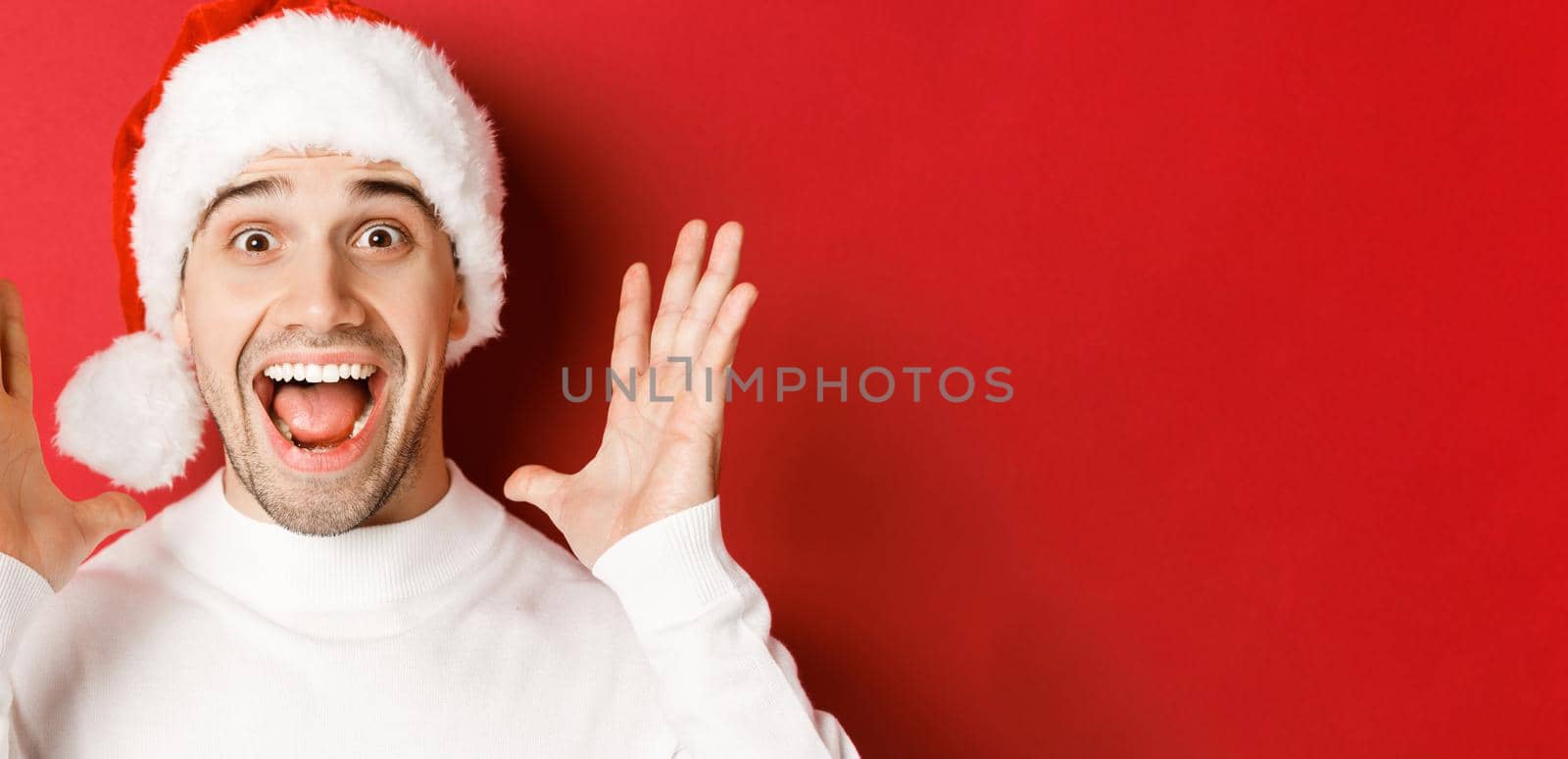 Close-up of happy young man in santa hat, making big christmas announcement, smiling amazed, standing over red background.