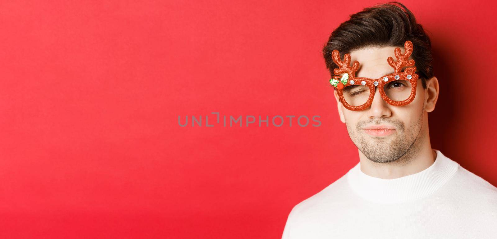 Concept of winter holidays, christmas and celebration. Close-up of cheeky attractive man in party glasses, winking and looking sassy, standing over red background.