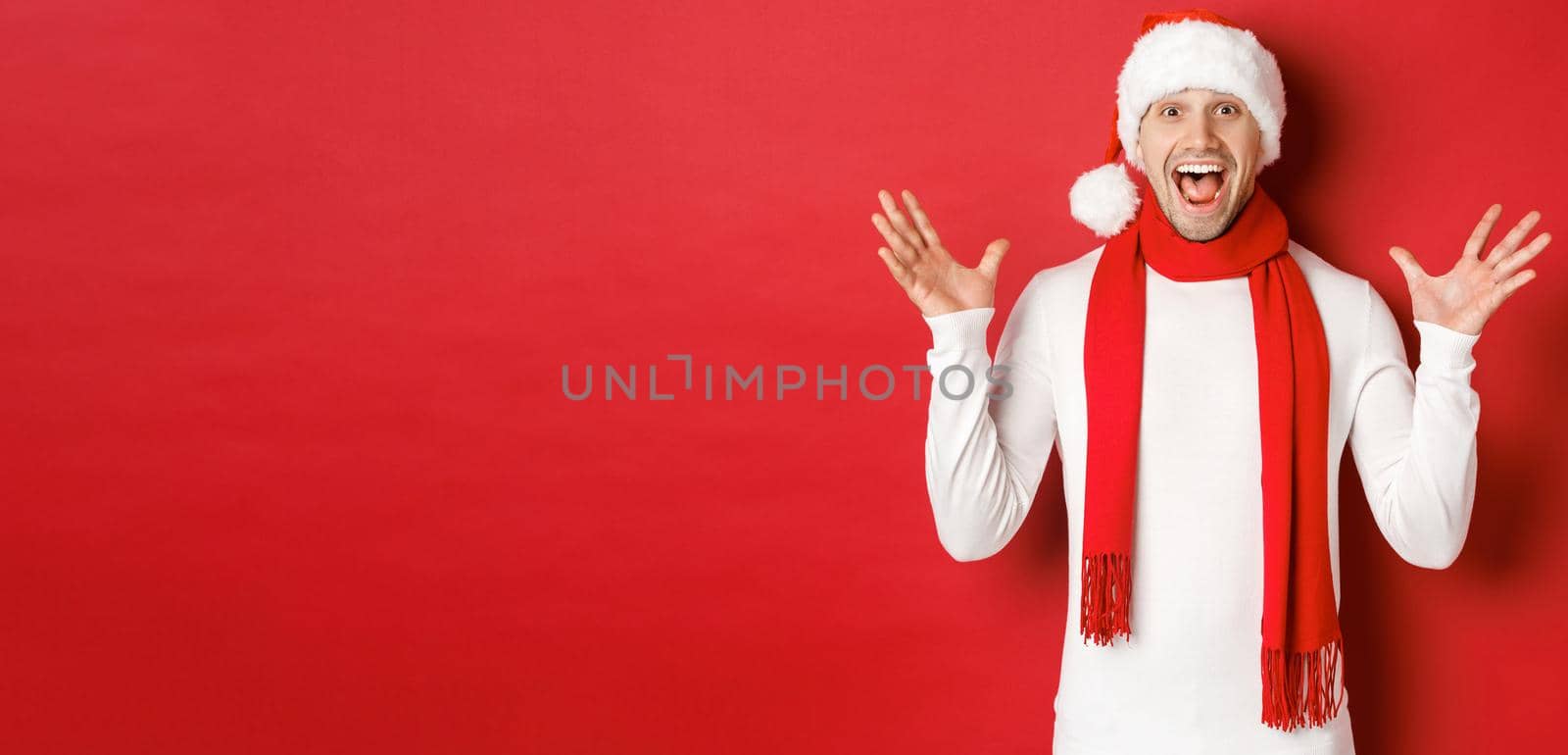 Portrait of happy and amazed handsome man, celebrating new year, wishing merry christmas, wearing santa hat and scarf, telling big news, standing against red background by Benzoix