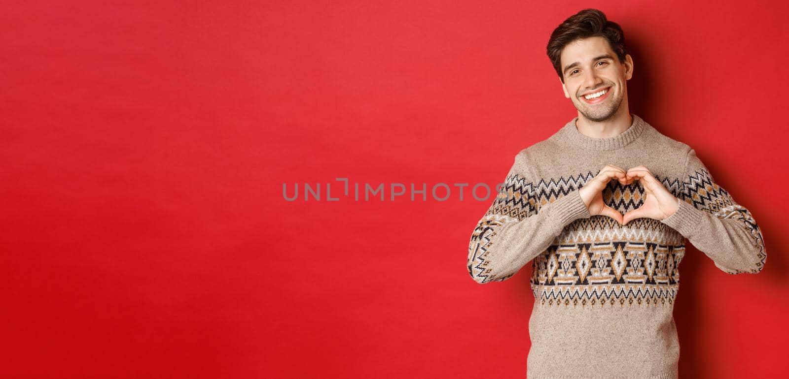 Image of handsome bearded guy in winter sweater, wishing merry christmas and showing heart sign, smiling lovely at camera, standing against red background by Benzoix