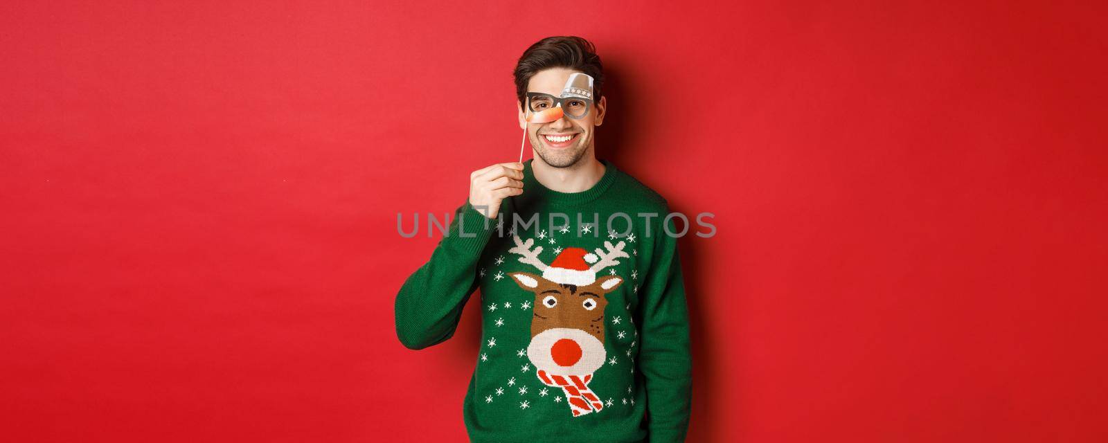 Funny man in christmas sweater and party mask, celebrating winter holidays, smiling happy, standing over red background.