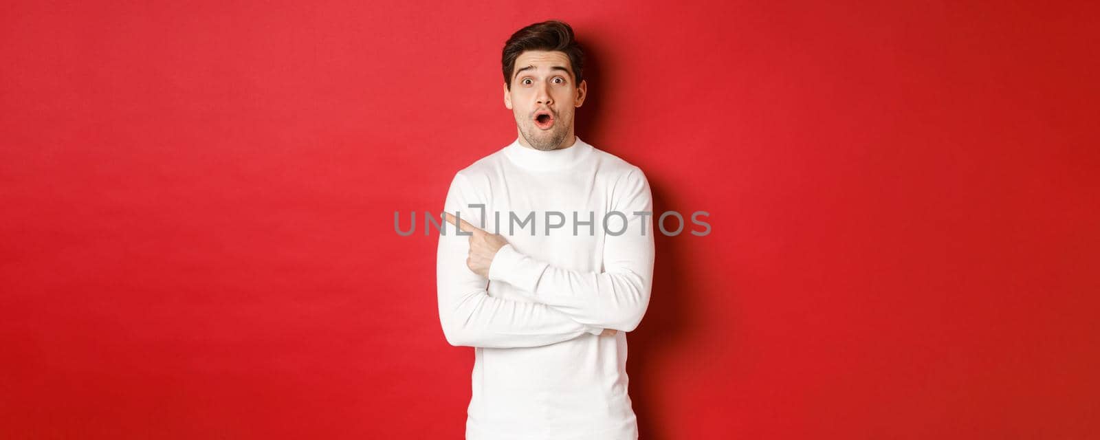 Concept of winter holidays, christmas and lifestyle. Surprised handsome guy pointing finger at new year advertisement, pointing finger left and drop jaw amazed, standing over red background by Benzoix