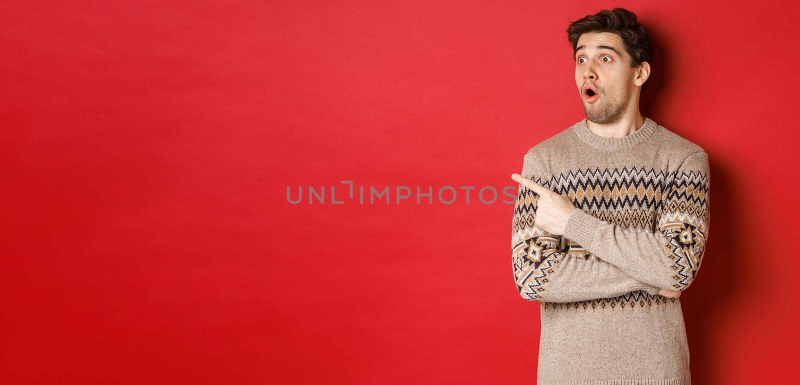 Portrait of attractive caucasian man celebrating new year, winter holidays, pointing finger and looking left surprised, showing christmas advertisement, standing over red background.
