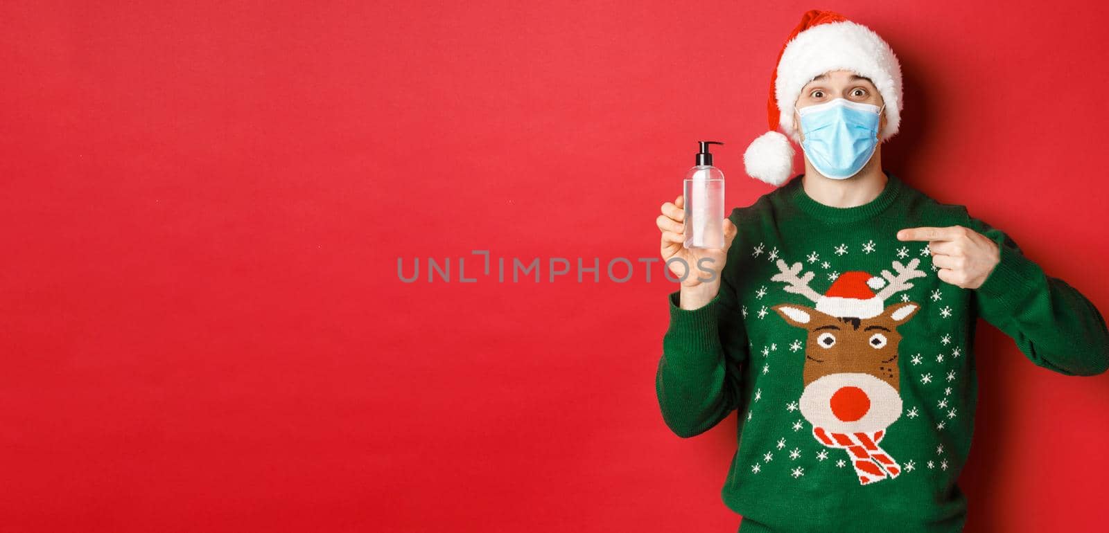 Concept of new year, coronavirus and social distancing. Portrait of cheerful man in santa hat, christmas sweater and medical mask, pointing finger at hand sanitizer, standing over red background.