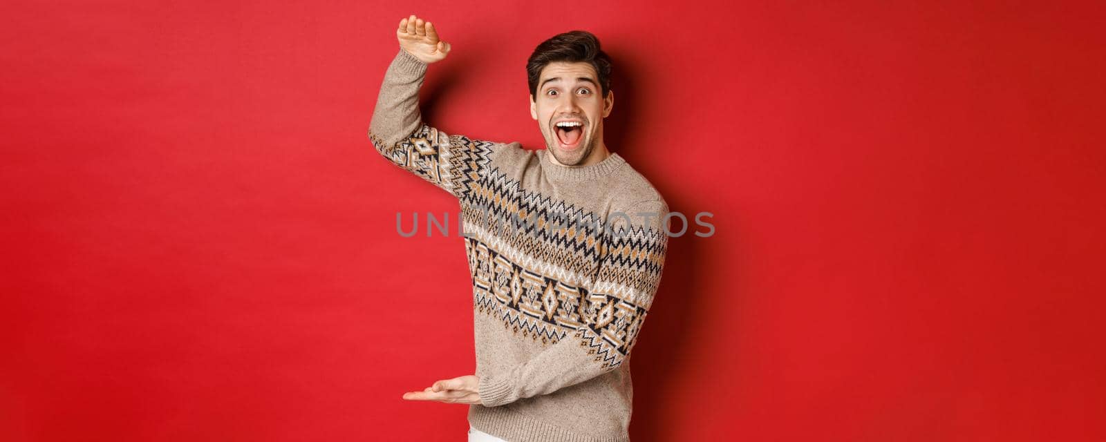 Portrait of excited and happy handsome man in christmas sweater, showing something big, holding large gift for holidays, standing over red background.