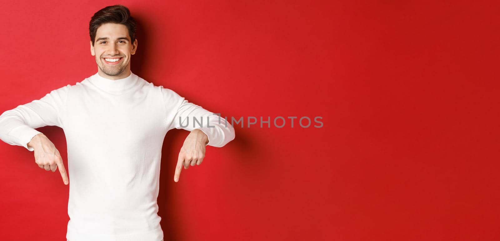 Image of smiling handsome man in white sweater inviting visit page, pointing fingers down and showing christmas banner, standing against red background by Benzoix