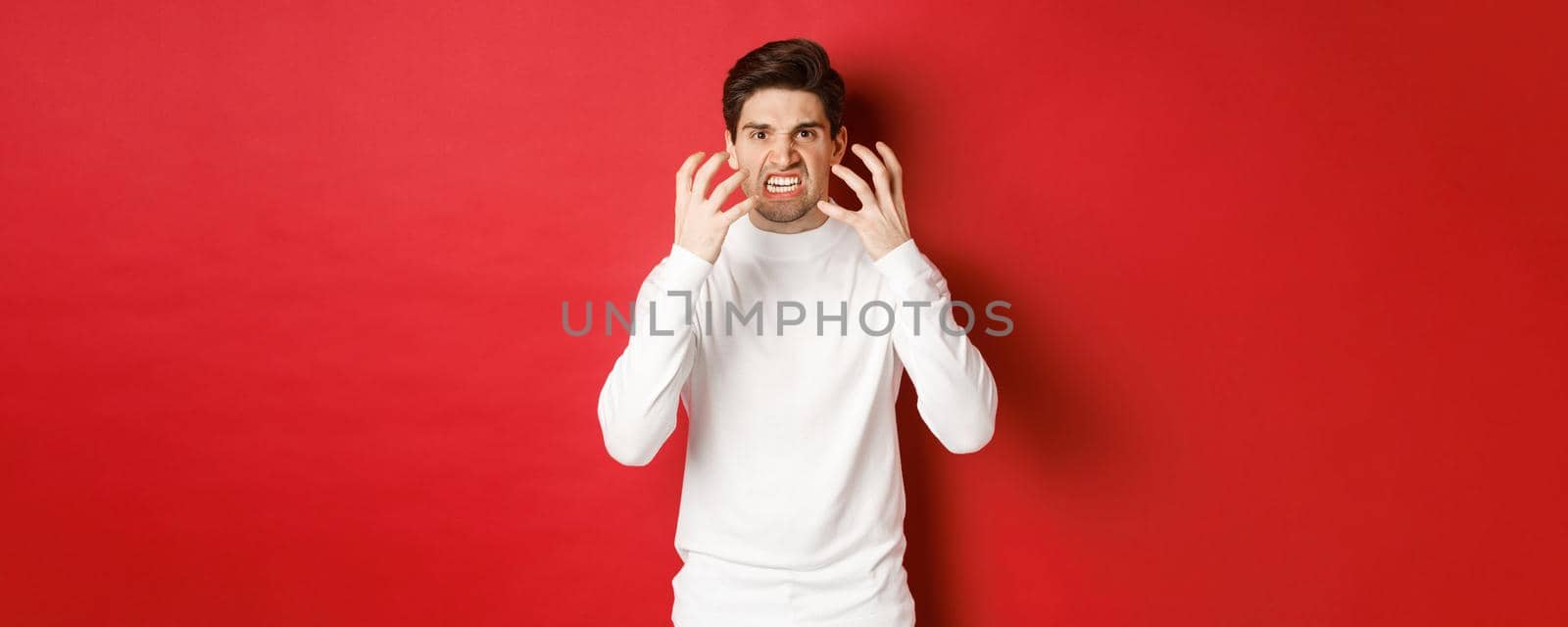 Image of angry and pissed-off man in white sweater, grimacing and shaking from rage, standing furious against red background.