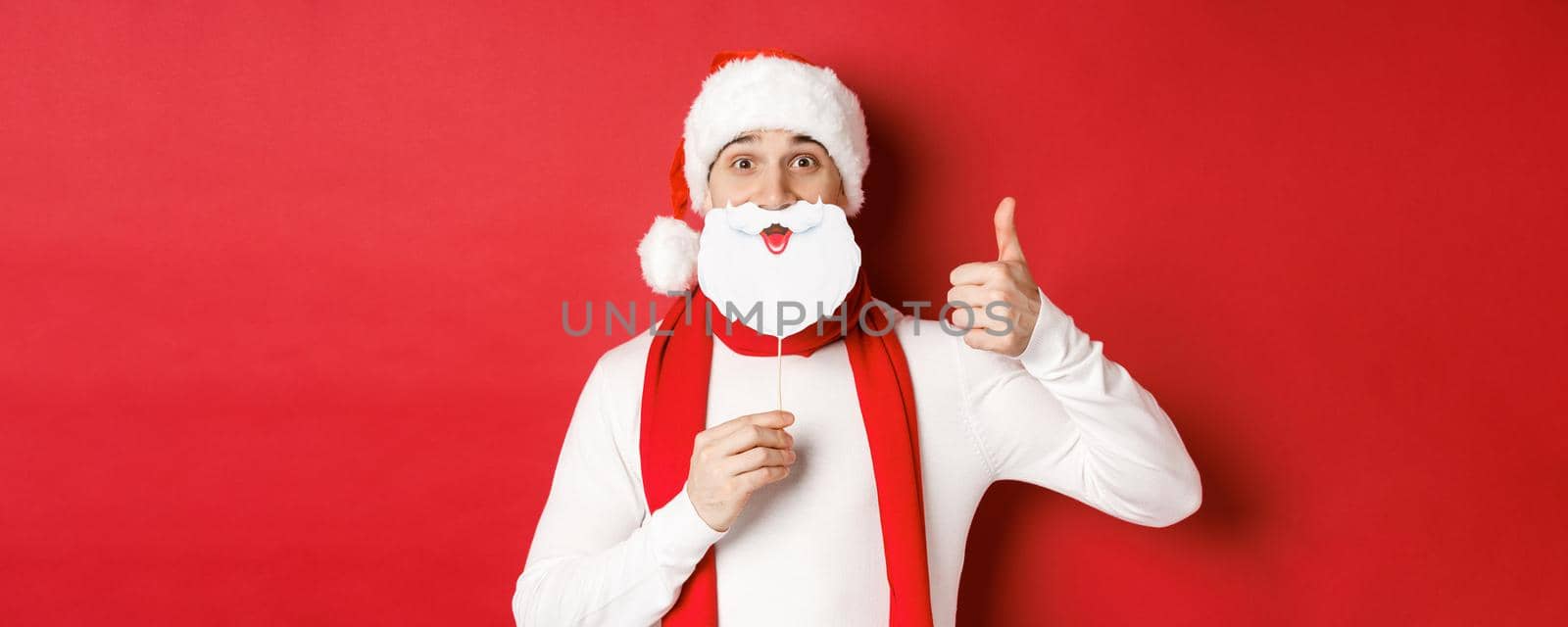 Concept of christmas, winter holidays and celebration. Portrait of funny man in santa hat, holding beard mask, showing thumb-up, enjoying new year party, standing over red background by Benzoix
