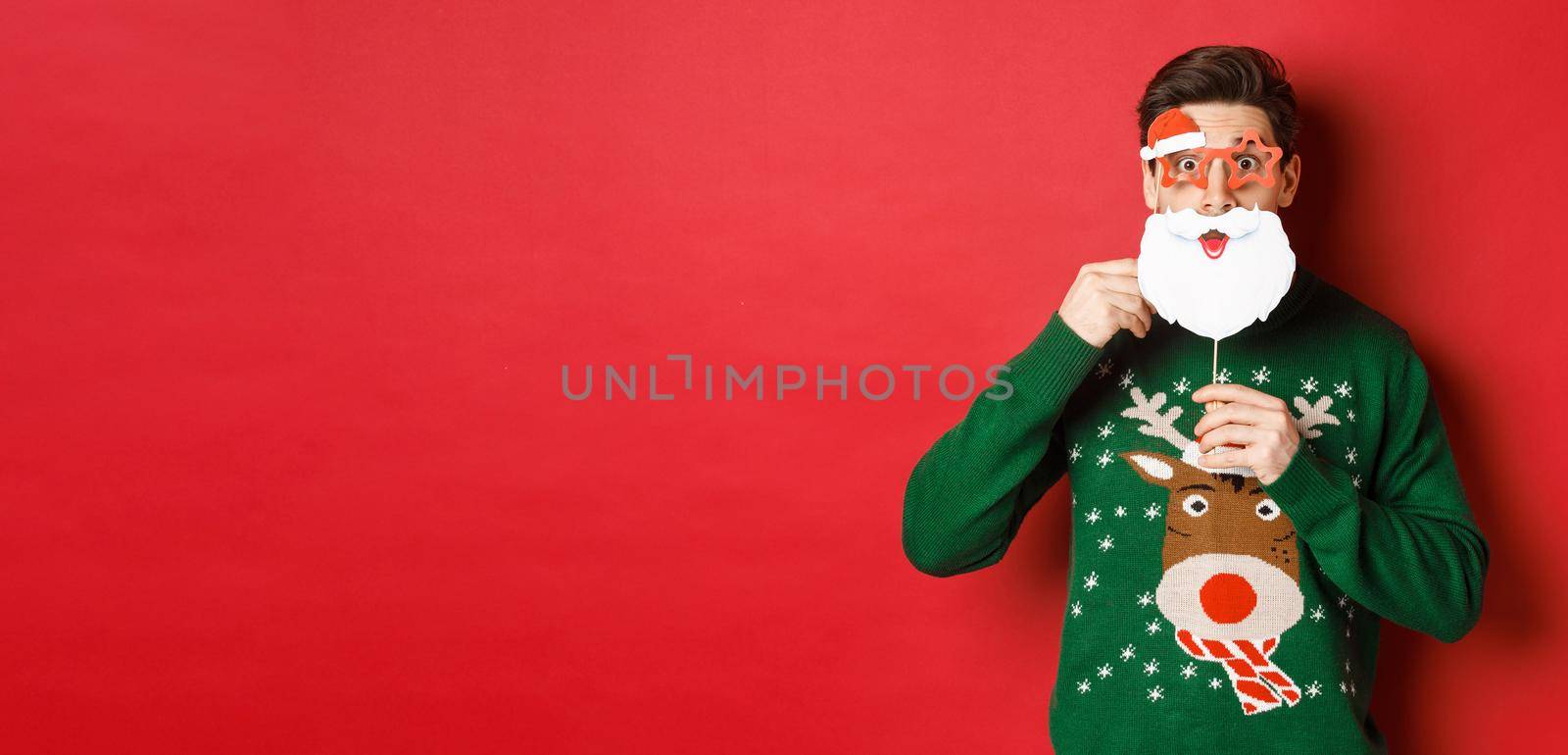Portrait of surprised man in green christmas sweater, holding funny santa claus mask, celebrating new year, standing over red background.