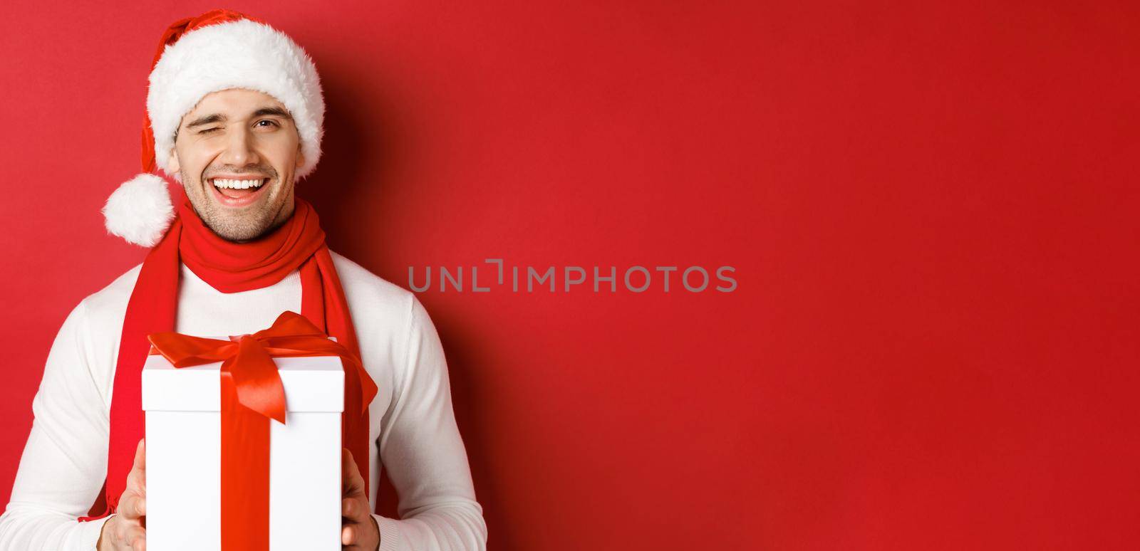 Concept of winter holidays, christmas and lifestyle. Handsome cheeky man in santa hat and scarf, holding present and smiling, winking at camera, standing over red background.