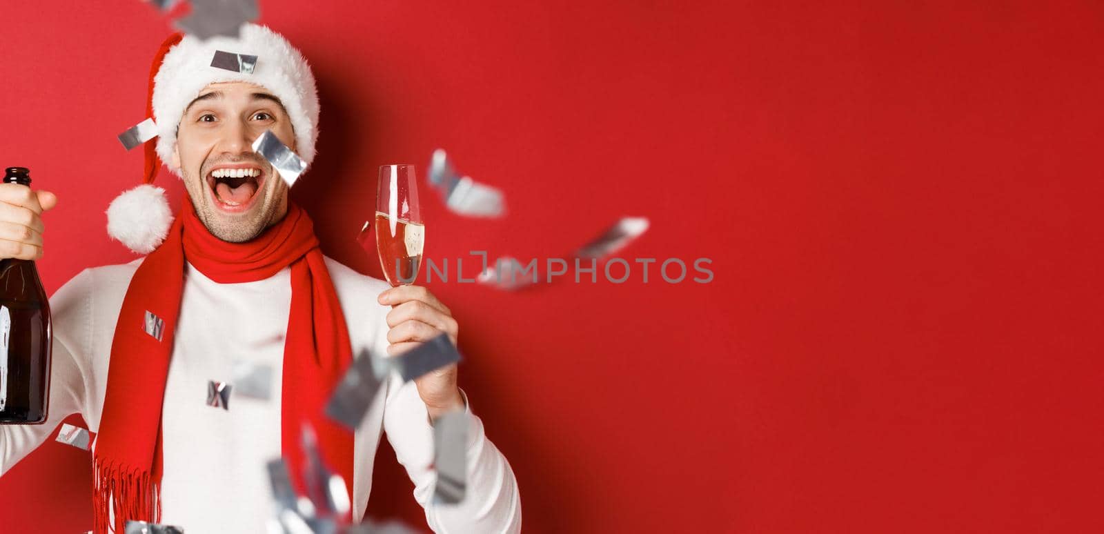 Concept of winter holidays, christmas and lifestyle. Close-up of happy man celebrating new year, holding champagne bottle and glass, standing over red background with confetti by Benzoix