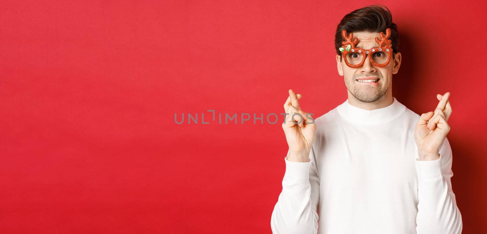 Close-up of handsome nervous guy in party glasses, making a wish, crossing fingers for good luck and looking with hope at camera, standing over red background by Benzoix