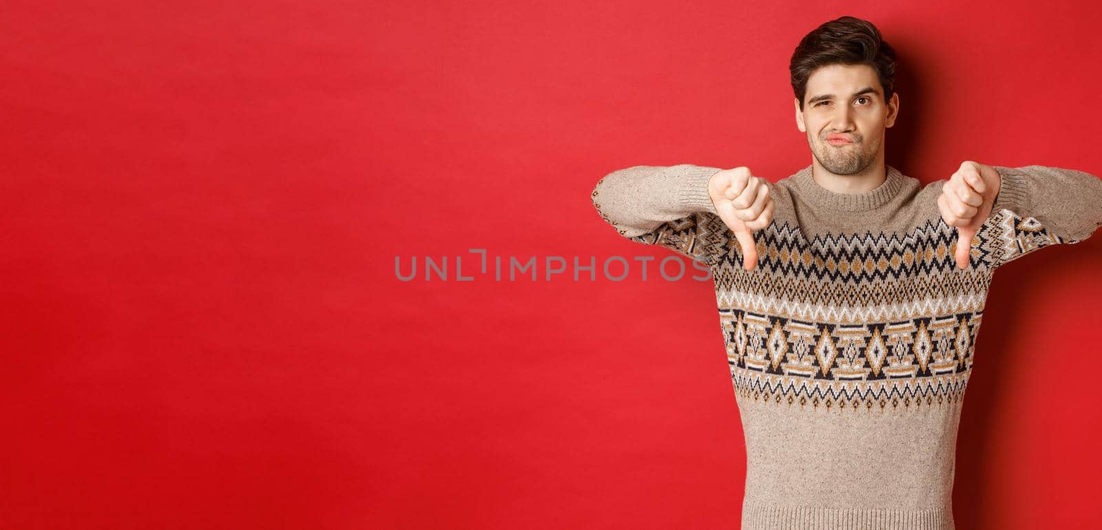 Portrait of skeptical and disappointed handsome man in christmas sweater, dislike new year party, showing thumbs-down, express disapproval, standing over red background by Benzoix