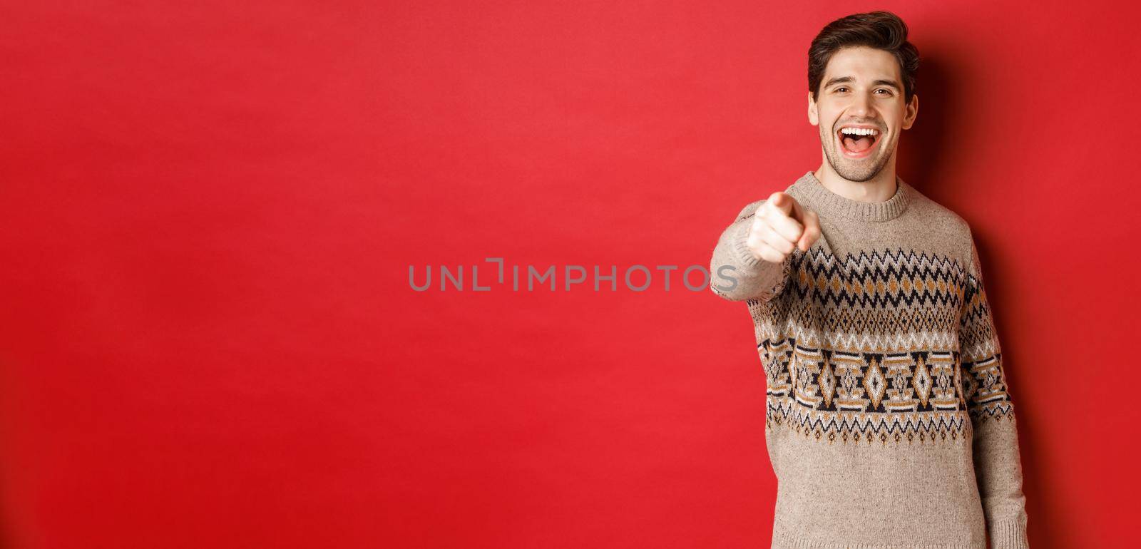 Concept of christmas celebration, winter and lifestyle. Happy attractive man in xmas sweater, wishing happy holidays and pointing at camera, standing over red background by Benzoix