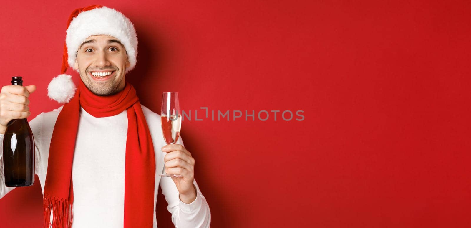 Concept of winter holidays, christmas and lifestyle. Close-up of cheerful handsome man, holding champagne bottle and glass, making toast for new year and celebrating, red background by Benzoix