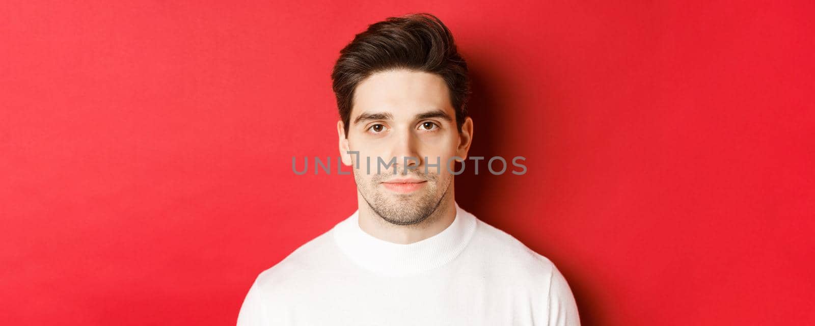Close-up of handsome brunette guy in white sweater, smiling at camera, standing over red background by Benzoix