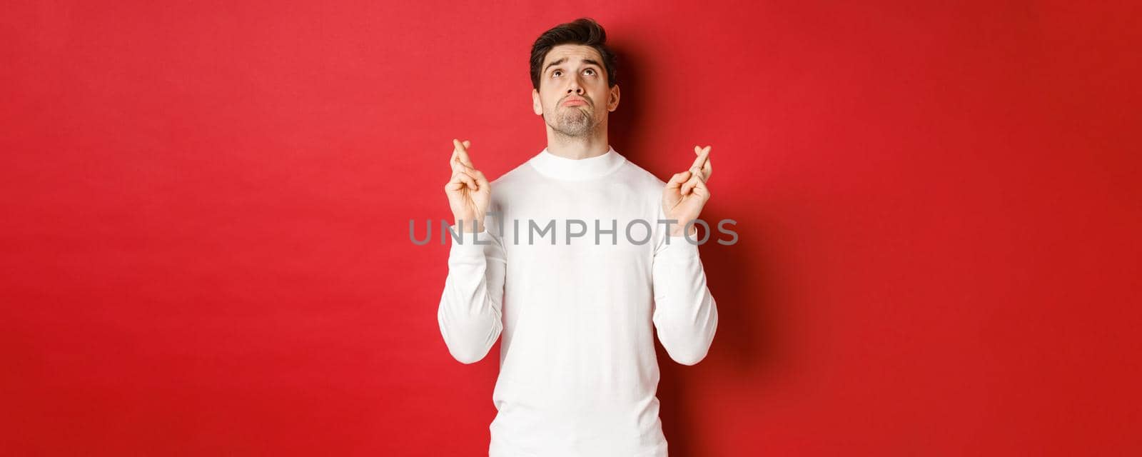Portrait of sad and hopeful man making a wish, begging god and looking up, crossing fingers for good luck, standing over red background by Benzoix
