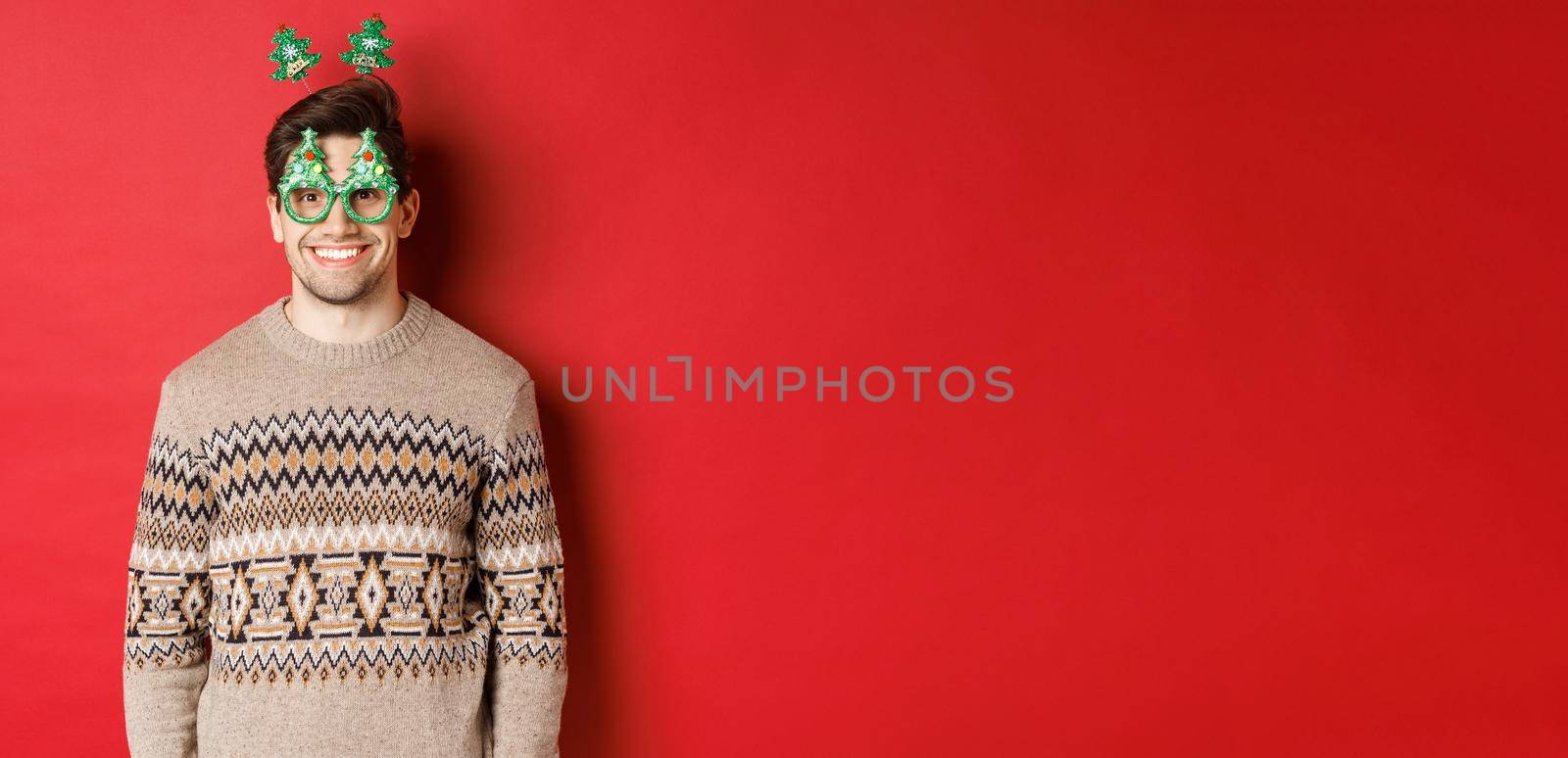 Portrait of happy and cute man in winter sweater and party glasses, celebrating new year or christmas, smiling while standing over red background.