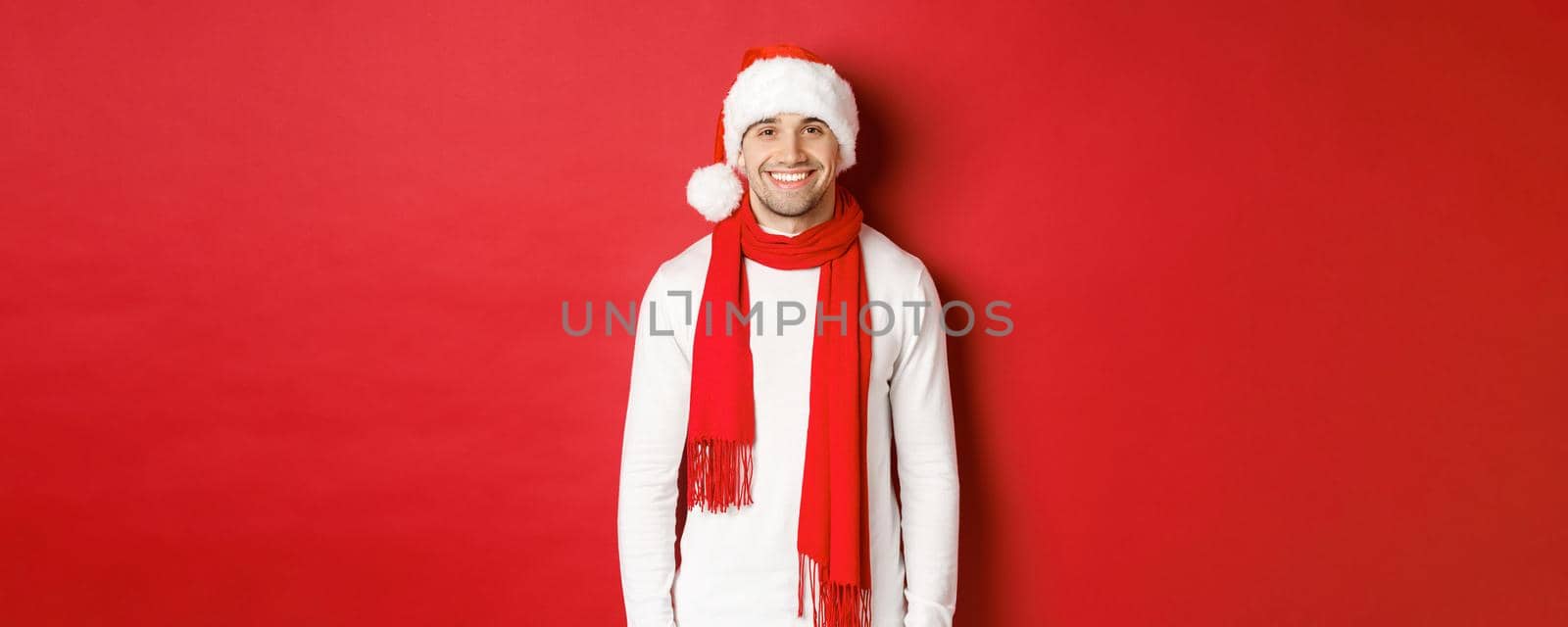 Happy caucasian guy celebrating christmas and smiling, wearing santa hat, scarf and white sweater, enjoying new year holidays, standing over red background.