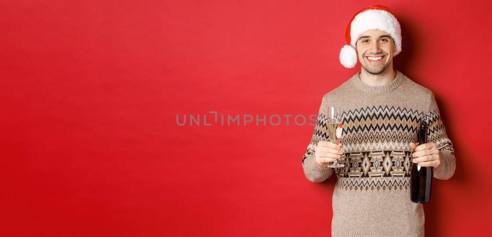 Concept of winter holidays, new year and celebration. Portrait of handsome man in santa hat and sweater, holding champagne and making toast on christmas party, standing over red background.