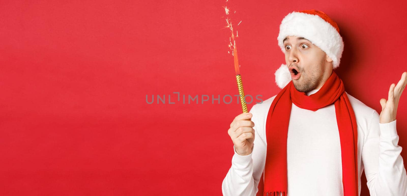 Concept of christmas, winter holidays and celebration. Close-up of amazed handsome man in santa hat and scarf, looking at sparkler with excitement, standing over red background.